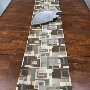 May include: A rectangular table runner featuring a geometric pattern of overlapping squares in shades of brown, beige, and grey. Resting on top of the runner is a silver-toned, fish-shaped decorative dish with a textured, scale-like surface finish.