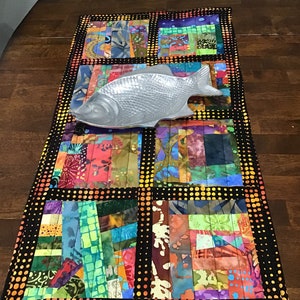 May include: A colorful patchwork table runner with a black background and a polka dot border. The runner is made of different fabrics with a variety of colors and patterns. A silver fish-shaped serving dish is placed on top of the runner.