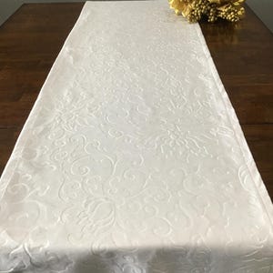 May include: A white table runner with an embossed floral pattern. The runner is rectangular and lies on a dark wooden table. A gold floral decoration sits at the top right corner of the table.