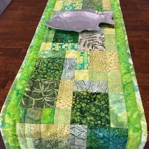 May include: A rectangular quilted table runner featuring a patchwork of various green and yellow fabric squares with diverse patterns, including leaves, circles, and abstract lines. A silver-toned metal fish-shaped dish rests on top.