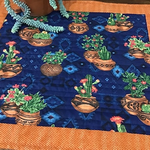 May include: A blue quilted table runner with a southwestern design featuring multiple potted cacti and succulents. The runner has an orange border.