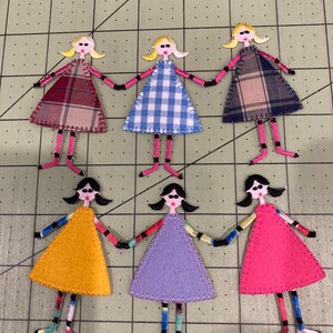 May include: Six handmade fabric doll figures. The dolls have blonde or black hair, sunglasses, and are wearing dresses in various colors and patterns, including plaid and gingham. The dolls are connected by their hands.