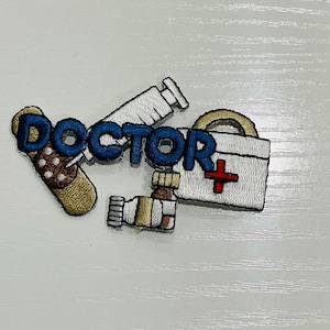 May include: An embroidered patch featuring the word DOCTOR in blue letters, accompanied by a white syringe, a tan adhesive bandage with brown dots, a small brown medicine bottle, and a white medical bag with a red cross symbol.