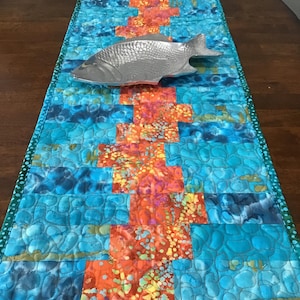 May include: A quilted table runner with a vibrant blue and orange pattern. The runner features a central design of orange blocks with a colorful, speckled pattern, set against a blue background. A silver fish-shaped dish rests on top.