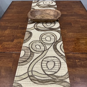 May include: A decorative table runner with a repeating pattern of brown and tan circles. A wooden bowl sits atop the runner. The runner is placed on a dark brown wooden table.