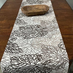 May include: A rectangular table runner featuring a repeating pattern of dark brown and light beige leaf silhouettes on a cream-colored fabric base. A small, oval-shaped, dark brown wooden bowl with a smooth, carved texture sits on the runner.