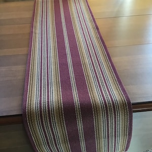 May include: A striped table runner with burgundy and beige stripes. The runner is made of a woven fabric and has a fringed edge.