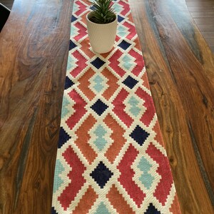 May include: A colorful table runner with a geometric diamond pattern in shades of red, orange, blue, and white. The runner has a fringed edge and is laid on a wooden table.
