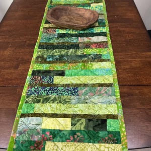 May include: A rectangular quilted table runner featuring a patchwork of various green, lime, and olive fabric strips with diverse patterns. A small, oval-shaped, dark brown wooden bowl rests on the center of the runner, which has a lime green border.