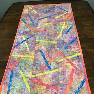 May include: A rectangular, quilted table runner with a colorful abstract design. The background features a mix of colors, with intersecting lines in blue, orange, and yellow. The edges are finished with a pink border.
