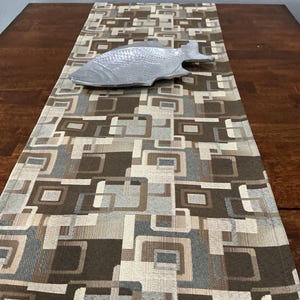 May include: A rectangular table runner featuring a geometric pattern of overlapping squares in shades of brown, beige, and grey. Resting on the runner is a silver-toned metal decorative dish shaped like a fish with a textured scale pattern.