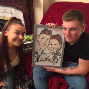May include: A framed caricature drawing of a couple in a black frame. The couple is depicted in a cartoon style with exaggerated features. The man is wearing a blue shirt and the woman is wearing a black dress. The drawing is set against a white background.