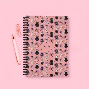 Op de afbeelding: Een roze notitieboek met een zwarte spiraalbinding, versierd met een herhalend patroon van zwarte katten, drankflessen, handen en spreukenboeken. Het woord "NOTES" staat op de kaft. Een roze pen en een gouden paperclip zijn zichtbaar.