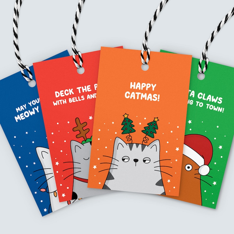 K&ouml;nnte beinhalten: Vier farbige Weihnachtsanh&auml;nger mit Katzenmotiven und festlichen Spr&uuml;chen. Die Anh&auml;nger sind in Blau, Rot, Orange und Gr&uuml;n gehalten und tragen Slogans wie "May You Meowy" und "Happy Catmas!". Jedes Etikett ist mit schwarz-wei&szlig;em Garn versehen.