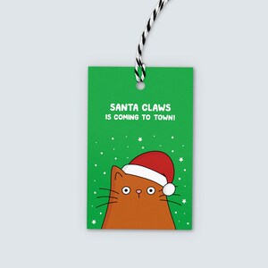 K&ouml;nnte beinhalten: Ein gr&uuml;nes Geschenk-Etikett mit dem Text "SANTA CLAWS IS COMING TO TOWN!" zeigt eine Cartoon-Katze mit Weihnachtsm&uuml;tze. Die Katze ist braun mit einem rot-wei&szlig; umrandeten Hut. Das Etikett hat eine schwarz-wei&szlig;e Schnur.