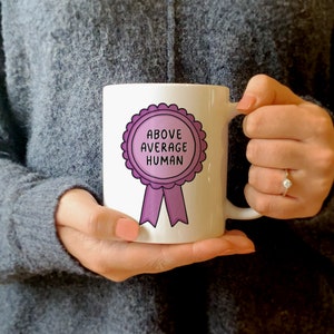 Könnte beinhalten: Eine weiße Keramiktasse mit einem rosa Band und einem lila Kreis, auf dem "Above Average Human" steht.