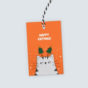 K&ouml;nnte beinhalten: Orangefarbenes Geschenk-Etikett mit dem Text "HAPPY CATMAS!" und einer Cartoon-Katze mit Weihnachtsbaum-Geweihen. Die Katze ist grau und wei&szlig; mit einem m&uuml;rrischen Gesichtsausdruck. Eine schwarz-wei&szlig;e Schnur ist befestigt.