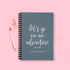 Può includere: Un quaderno a spirale grigio con il testo "Let's go on an adventure" in lettere bianche in corsivo. Il quaderno ha anche una piccola freccia bianca che punta a destra e il testo "Kate's Travel Notebook" in bianco.