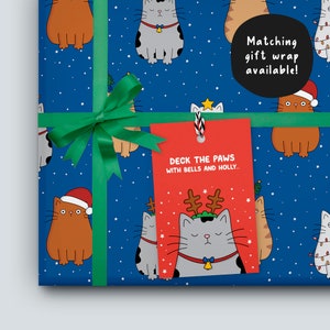 K&ouml;nnte beinhalten: Geschenkpapier mit blauem Hintergrund, Cartoon-Katzen und wei&szlig;en Sternen. Ein gr&uuml;nes Band und eine Schleife sichern die Verpackung. Ein rotes Etikett lautet: "DECK THE PAWS WITH BELLS AND HOLLY..." und "Matching gift wrap available!"