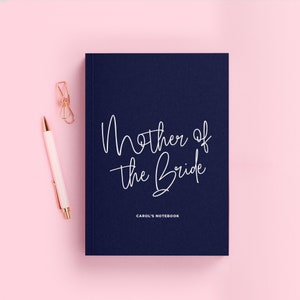 May include: A navy blue notebook with the text "Mother of the Bride" in white script. The notebook has the text "CAROL'S NOTEBOOK" printed in white at the bottom.
