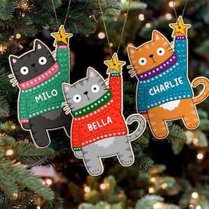 May include: Three Christmas tree decorations shaped like cats wearing jumpers. The cats are holding a star in their paw. The jumpers are green, red, and blue with white snowflakes. The cats are named Milo, Bella, and Charlie.