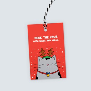 K&ouml;nnte beinhalten: Rotes Geschenk-Etikett mit einer Cartoon-Katze, die ein Rentiergeweih und einen Glockenhalsband tr&auml;gt. Der Text lautet: &bdquo;Deck the Paws with Bells and Holly...&ldquo; in Wei&szlig;. Das Etikett ist mit schwarz-wei&szlig;em Garn versehen.