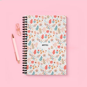 Op de afbeelding: Een wit notitieboek met spiraalbinding en een kleurrijk bloemenpatroon met bladeren, bloemen en eikels. De omslag heeft de tekst "NOTES" in zwart gedrukt.