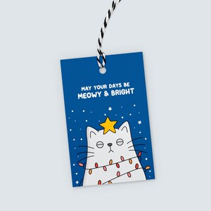 K&ouml;nnte beinhalten: Ein blaues Geschenk-Etikett mit dem Text "MAY YOUR DAYS BE MEOWY & BRIGHT". Das Etikett zeigt eine wei&szlig;e Katze mit einem Stern auf dem Kopf und Lichterketten. Ein schwarz-wei&szlig;es Band ist befestigt.