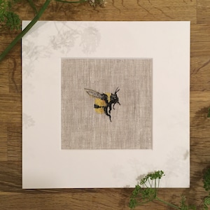 Può includere: Opera d'arte ricamata raffigurante un calabrone in volo, con strisce gialle e nere e ali dettagliate. L'ape è posta su uno sfondo beige con texture di lino, incorniciata da un passe-partout bianco. L'opera è presentata su una superficie di legno con rametti verdi.