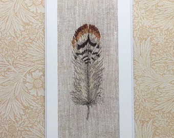 May include: A single brown and white feather with a brown tip is displayed on a textured linen background. The feather is centered on the card and is the main focus of the image.