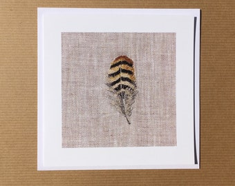 May include: A square greeting card featuring an embroidered feather design. The feather has brown, black, and tan stripes with a detailed, feathery base. The card is set against a neutral linen-textured background, framed by a white border.