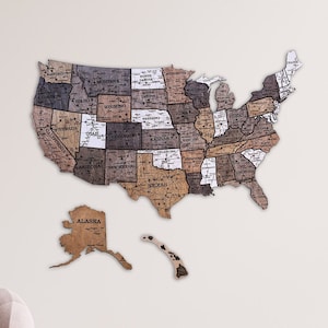 May include: A wooden map of the United States with each state outlined and labelled. The map is mounted on a white wall in a living room setting. The map is a light brown colour with darker brown accents.