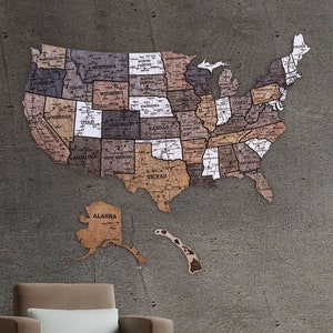 May include: A wooden map of the United States with each state outlined and labelled. The map is mounted on a grey wall with a brown leather armchair in the foreground.