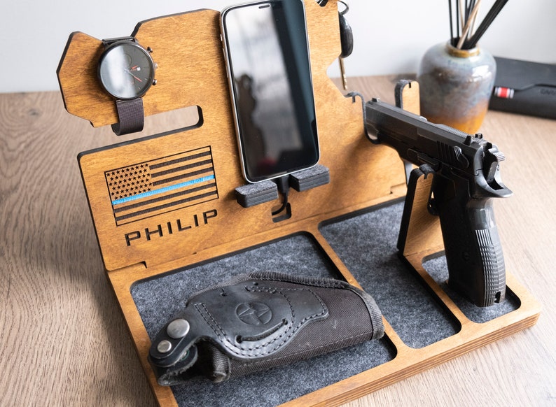 Puede incluir: Organizador de madera con compartimentos para un tel&eacute;fono, un reloj y una pistola. Presenta una bandera estadounidense con una l&iacute;nea azul y el nombre "PHILIP" grabado en ella.