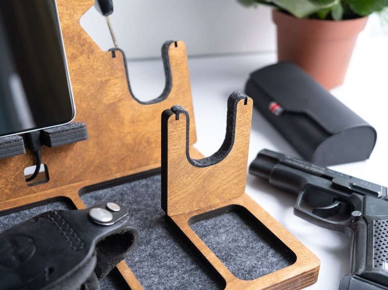 Puede incluir: Organizador de madera con m&uacute;ltiples compartimentos para guardar diversos art&iacute;culos. Cuenta con un soporte para tel&eacute;fono, un soporte para reloj, un soporte para pistola y una bandeja peque&ntilde;a. El organizador est&aacute; hecho de madera marr&oacute;n claro y tiene un forro de fieltro gris.