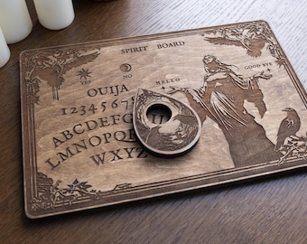 May include: A wooden spirit board with engraved letters, numbers, and symbols. The board features a detailed illustration of a woman, ravens, and other decorative elements. A teardrop-shaped planchette rests on the board.