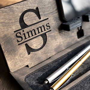 May include: A wooden desk organiser with a personalised monogram of the letter "S" and the name "Simms". The organiser has a slot for a phone, a pen holder, and a small compartment for other items.