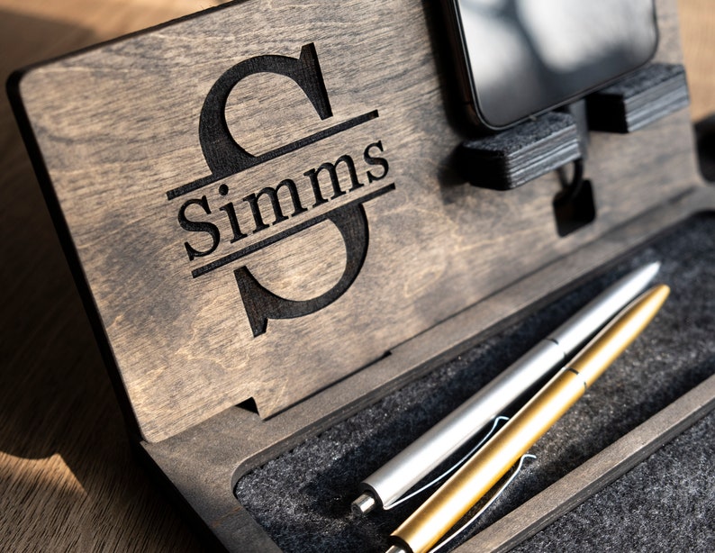 May include: A wooden desk organiser with a personalised monogram of the letter "S" and the name "Simms". The organiser has a slot for a phone, a pen holder, and a small compartment for other items.