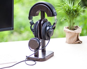 Puede incluir: Un soporte para auriculares de madera marrón oscuro con una parte superior curva y una base. Un par de auriculares negros están apoyados en el soporte. El soporte está sobre una superficie blanca.