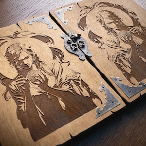 Puede incluir: Caja de madera con un diseño detallado de ángel cortado con láser en cada lado. La caja es de color marrón claro con detalles plateados en las esquinas y un cierre de metal. La obra de arte presenta detalles intrincados y un halo.