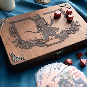 Könnte beinhalten: Eine Holzkiste mit einem lasergravierten Design eines Bogenschützen, umgeben von dekorativen Eckakzenten. Rote Würfel und ein Kartenspiel liegen in der Nähe der Kiste. Die Kiste steht auf einer blauen Oberfläche, mit Kerzen und Münzen im Hintergrund.