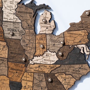 May include: A wooden map of the United States with push pins marking different states. The map is made of different shades of brown wood and has a rustic look.