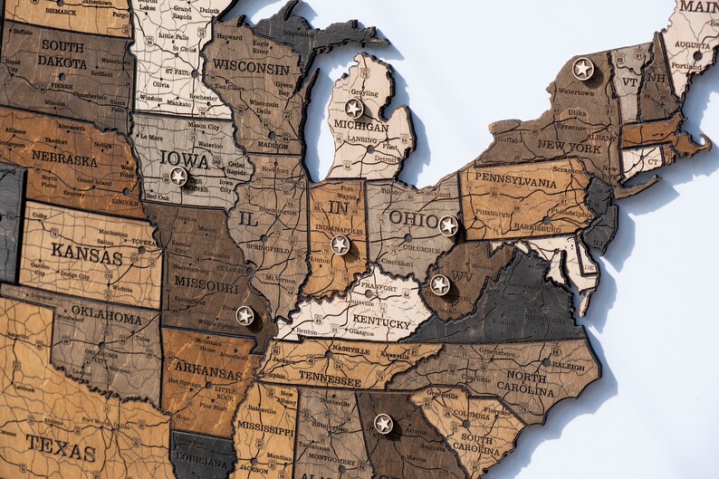 May include: A wooden map of the United States with push pins marking different states. The map is made of different shades of brown wood and has a rustic look.