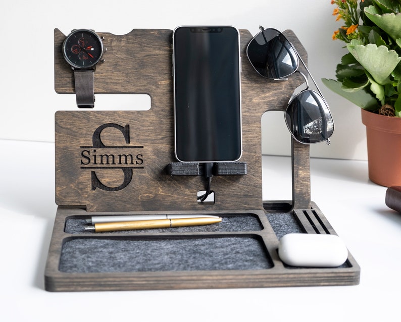 May include: A wooden organiser with a grey felt lining. It has slots for a phone, watch, sunglasses, and pens. The organiser is personalised with the name "Simms".