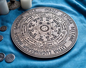 May include: A silver-colored, circular decorative plate with engraved text and symbols. The plate features a central sun design surrounded by astrological symbols and text indicating seasonal events like Samhain, Yule, and Litha. The plate is on a blue surface.