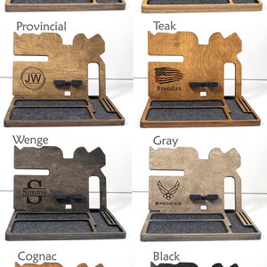 May include: Eight different wooden phone stands with various wood finishes and personalised engravings. The stands feature a phone holder, a watch holder, and a small tray. The engravings include names, initials, and symbols such as a fishing lure, an American flag, and an Air Force logo.