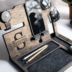 May include: A dark brown wooden organiser with a personalised name "Simms" engraved on the front. It has slots for a watch, phone, sunglasses, and pens. The organiser has a grey felt lining.