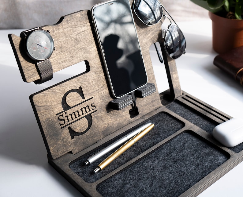 May include: A dark brown wooden organiser with a personalised name "Simms" engraved on the front. It has slots for a watch, phone, sunglasses, and pens. The organiser has a grey felt lining.