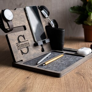 May include: A wooden organiser with a grey felt lining. It has slots for a phone, watch, and glasses. There is a small compartment for a wallet or other items. The organiser is personalised with the name "Simms".