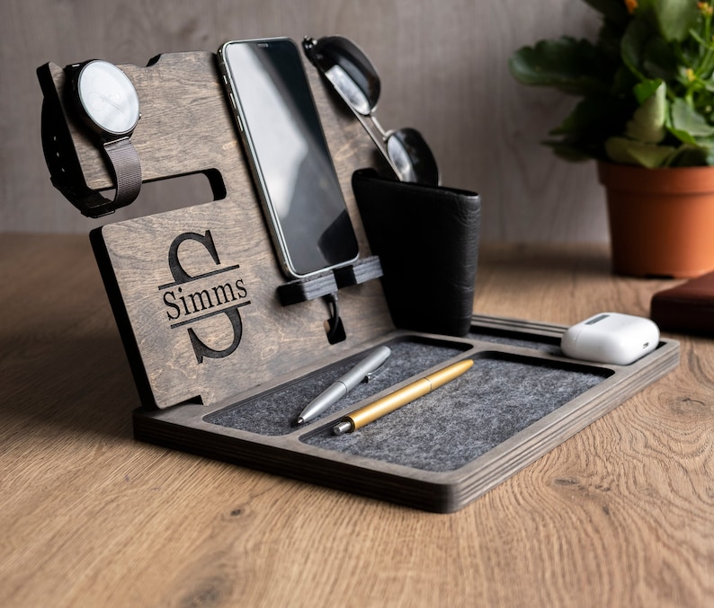 May include: A wooden organiser with a grey felt lining. It has slots for a phone, watch, and glasses. There is a small compartment for a wallet or other items. The organiser is personalised with the name "Simms".